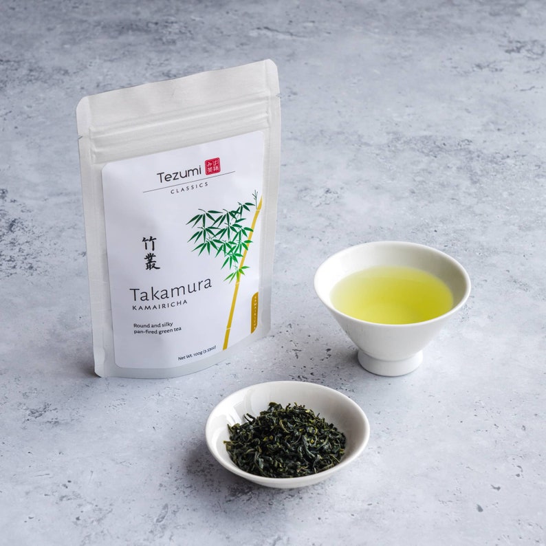 Puede incluir: Una bolsa blanca de t&eacute; Kamairicha "Tezumi Classics" con ilustraci&oacute;n de bamb&uacute;, un cuenco blanco peque&ntilde;o con t&eacute; suelto y una taza blanca llena de t&eacute; verde. El texto de la bolsa dice "Takamura" y "Round and silky pan-fired green tea."
