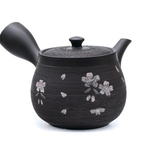 May include: Black ceramic teapot with a floral design. The teapot has a spout and a handle. The handle is shaped like a curved spout.