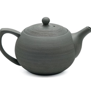 Puede incluir: Una tetera de cerámica gris oscuro con cuerpo redondeado y tapa. La tetera tiene un pico y una asa.