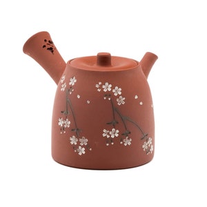 May include: A terracotta teapot with a floral design. The teapot has a small spout and a lid with a knob. The design features white flowers and green stems on a reddish-brown background.