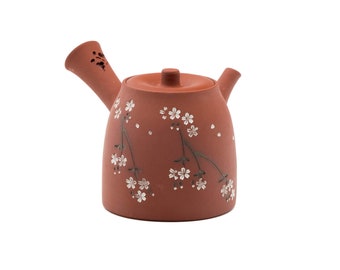 May include: A terracotta teapot with a floral design. The teapot has a small spout and a lid with a knob. The design features white flowers and green stems on a reddish-brown background.