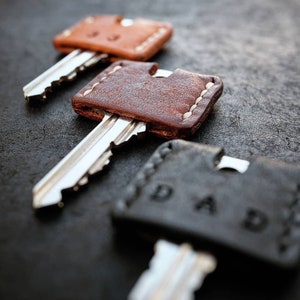 Op de afbeelding: Drie sleutels met leren sleutelhoezen. De hoezen zijn bruin, bruin en zwart. De zwarte hoes heeft het woord "DADDY" erop gegraveerd.