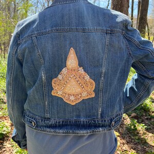 May include: A blue denim jacket with a gold embroidered patch on the back. The patch features a geometric design with floral accents.
