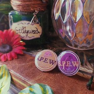 May include: Two round pins with a purple and white background. The pins feature a cartoon image of a creature with a purple body and white wings. The text on the pins reads "S.P.E.W. Proud Member".