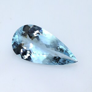 May include: A pear-shaped, light blue gemstone with facets. The gem has a translucent quality, with variations in color and internal reflections. The stone is set against a white background, highlighting its clarity and color.
