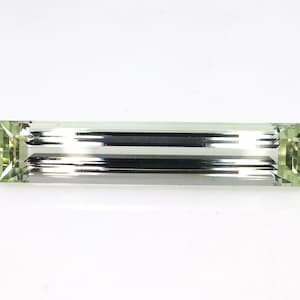 May include: A rectangular baguette-cut gemstone with a pale light green hue. The stone features long, parallel facets running along its length, creating a clear, reflective surface with sharp, defined edges and a transparent, crystalline appearance.