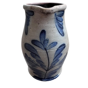 Puede incluir: Jarra de gres con base gris claro y estampado de hojas azul oscuro. La jarra tiene un borde ligeramente acampanado y un cuerpo bulboso. El diseño de hojas azules se repite. La jarra mide aproximadamente 20 cm de alto.