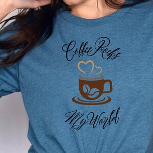 以下が含まれることがあります： 黒の筆記体で「Coffee Rocks My World」と書かれたターコイズブルーのTシャツ。テキストの上に、蒸気と2つのハートがついた茶色のコーヒーカップがあります。