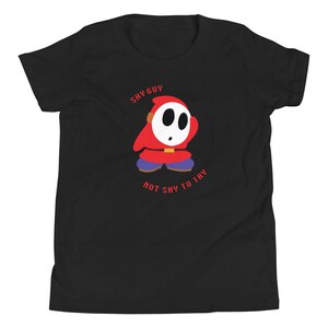 May include: Black youth t-shirt with a red and white Shy Guy character from the Super Mario video game series. The text "Shy Guy Not Shy To Try" is printed on the shirt.