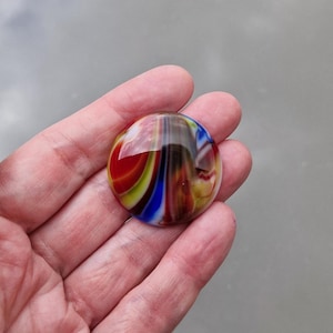 Könnte beinhalten: Ein kreisförmiger Glascabochon mit einer glatten, polierten Oberfläche und wirbelnden Mustern aus leuchtendem Rot, tiefem Blau, hellem Gelb und Weiß, die in einem marmorierten Effekt vermischt sind und ein glänzendes, reflektierendes Aussehen erzeugen.