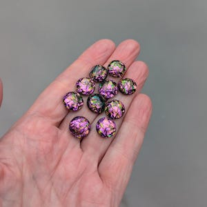 Puede incluir: Un conjunto de cuentas redondas de vidrio iridiscente en tonos morados, verdes y dorados. Las cuentas tienen un aspecto texturizado y fracturado. Se sostienen en una mano sobre un fondo gris. Adecuado para la fabricación de joyas.