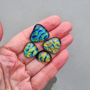 Puede incluir: Se muestran cuatro cabujones de vidrio dicróico en tonos azules, verdes y dorados. Los cabujones tienen un aspecto texturizado e iridiscente, con una forma redondeada y orgánica. Las piezas de vidrio se sostienen en una mano abierta sobre un fondo gris.