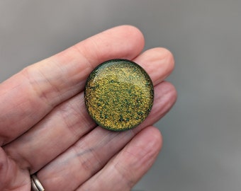 May include: A round, green and gold glitter resin cabochon. The cabochon is approximately 1 inch in diameter.