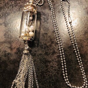 May include: A silver chain necklace with a glass bottle pendant filled with white pearls and a silver tassel. The bottle has a silver cap and a silver chain attached to the top.