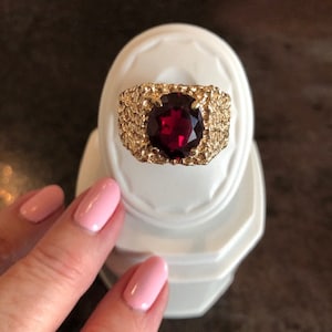 May include: A gold ring with a large, oval-shaped, dark red gemstone set in a textured band.