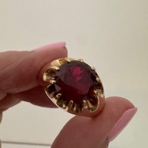 Op de afbeelding: Een goudkleurige ring met een grote, gefacetteerde, dieprode edelsteen. De zetting van de ring heeft een geschulpte vorm rond de edelsteen, wat een vintage of sierlijke uitstraling geeft. De facetten creëren een spel van licht en schaduw.