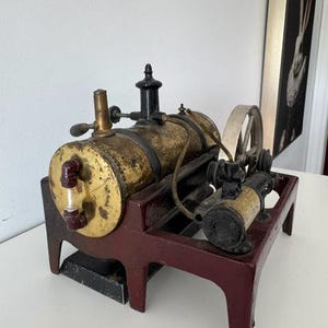May include: A vintage stationary steam engine model featuring a horizontal brass boiler mounted on a dark red cast iron base. The machine includes a black chimney, a silver flywheel, and a small pump mechanism connected by thin metal piping.