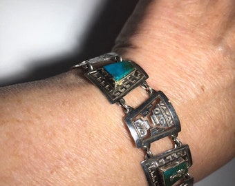Op de afbeelding: Een zilveren armband met turquoise stenen en ingewikkelde ontwerpen. De armband bestaat uit gekoppelde vierkanten met een gezichtsontwerp in elk vierkant.