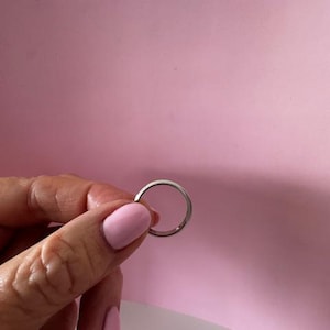 Pode incluir: Um anel de banda simples de prata segurado em uma mão com esmalte de unha rosa.