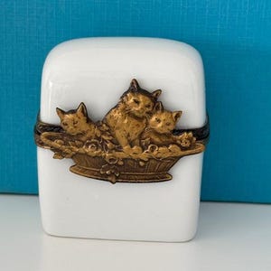 Könnte beinhalten: Eine weiße Porzellan-Schmuckdose mit einem goldfarbenen Metalldeckel, der drei Katzen in einem Korb mit Blumen zeigt.