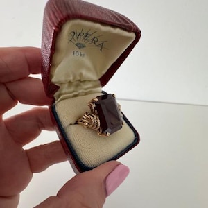 May include: A gold ring with a large, rectangular, dark red gemstone is displayed in a red jewellery box. The ring has an ornate, twisted gold band. The box is lined with cream-coloured fabric and has the word "PERA" and "10kt" printed on the inside.