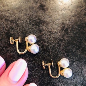 May include: Two pairs of gold-coloured clip-on earrings, each with two white pearls. The earrings are shown on a dark, textured surface. The image is a close-up shot.