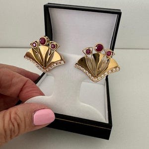 May include: A pair of gold earrings with a fan-shaped design. The earrings are adorned with red gemstones and clear diamonds.