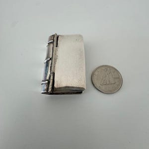 Può includere: Una scatola portaoggetti in argento a forma di libro con un coperchio incernierato. La scatola è chiusa e si trova su una superficie bianca accanto a una moneta d'argento.