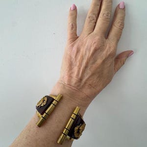 Puede incluir: Un brazalete de bisagra marrón y dorado con un diseño floral. El brazalete se lleva en la muñeca.