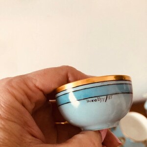 May include: A small, light blue teacup with a gold rim and a blue stripe. The cup has the inscription "Dorothy/53" on the bottom.