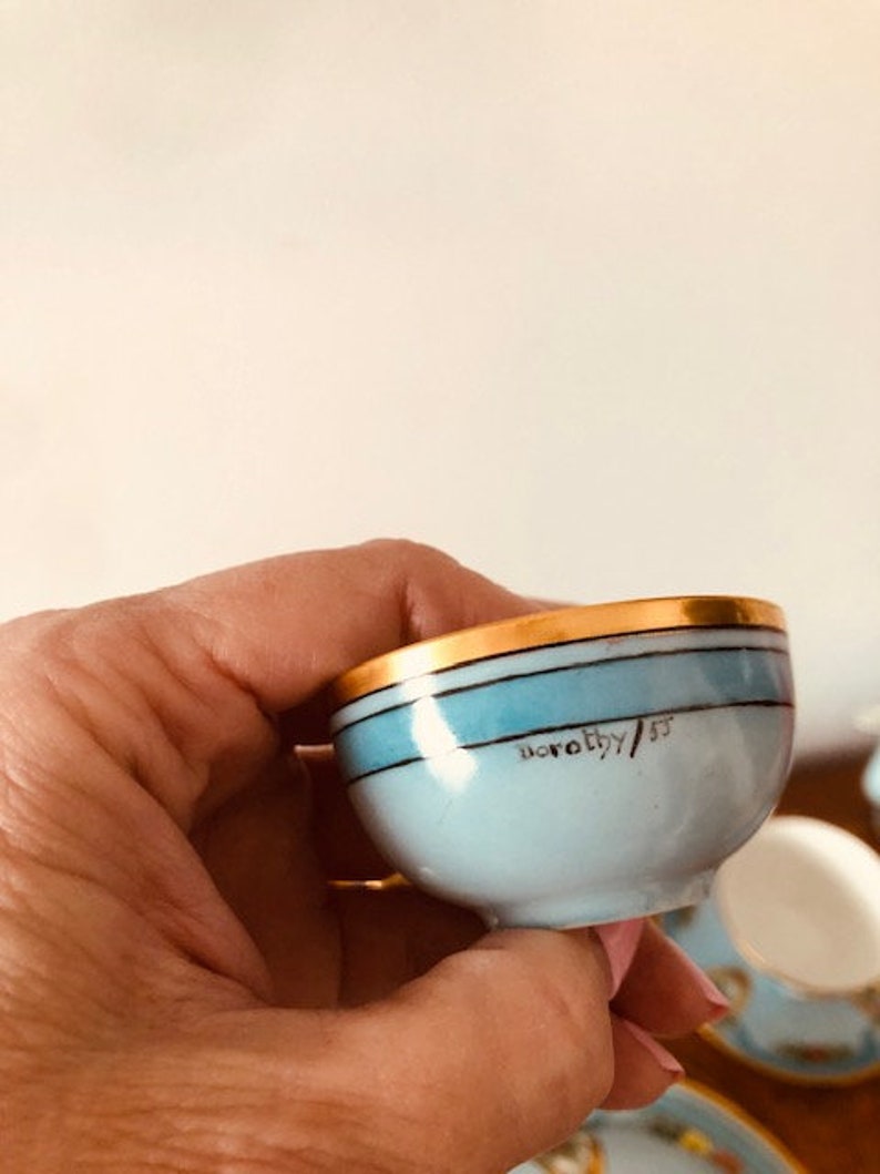 May include: A small, light blue teacup with a gold rim and a blue stripe. The cup has the inscription "Dorothy/53" on the bottom.