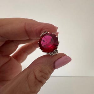 Peut inclure: Une bague en argent ornée d'une grosse pierre précieuse rouge rubis ronde. La pierre précieuse est le point central, avec une surface à facettes qui capte la lumière. Le corps de la bague présente un motif décoratif et texturé autour de la pierre.