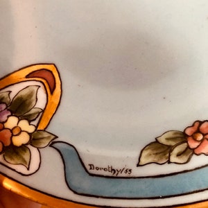 May include: A light blue ceramic plate with a gold rim and floral design. The plate has a blue ribbon with the text "Dorothy/55" on it.