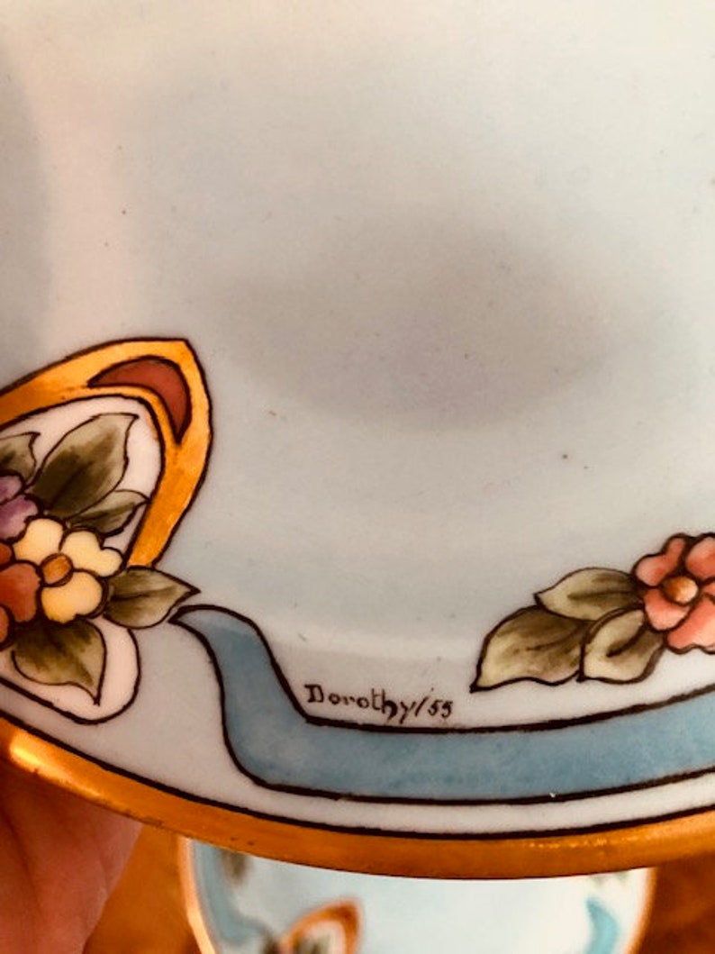 May include: A light blue ceramic plate with a gold rim and floral design. The plate has a blue ribbon with the text "Dorothy/55" on it.