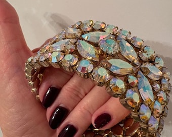 May include: A gold tone bracelet featuring iridescent rhinestones. The bracelet is made up of multiple rows of rhinestones and has a flexible design.