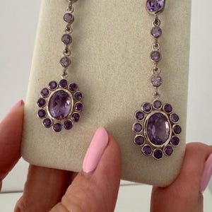 Può includere: Un paio di orecchini pendenti in argento con pietre preziose di ametista ovali e rotonde. Gli orecchini hanno un design floreale attorno alla grande ametista ovale nella parte inferiore. Gli orecchini sono esposti su una superficie beige.