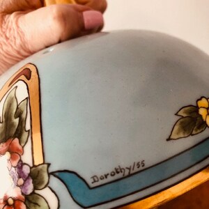 May include: A light blue ceramic serving dish with a gold rim and floral design. The dish has a lid with a gold knob and a blue ribbon with the inscription "Dorothy/55".
