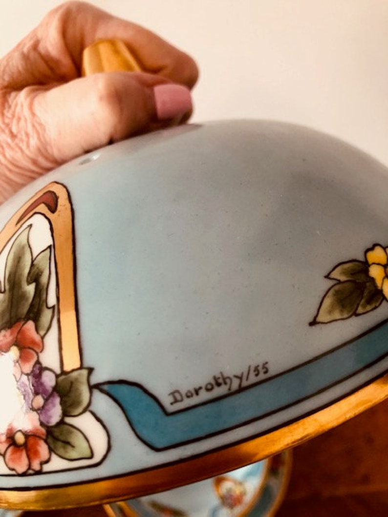 May include: A light blue ceramic serving dish with a gold rim and floral design. The dish has a lid with a gold knob and a blue ribbon with the inscription "Dorothy/55".