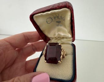 May include: A gold ring featuring a large, rectangular, dark red gemstone. The ring is presented in a red and blue jewellery box with the text "OPER" and "10k". The ring has a detailed, textured design on the band.