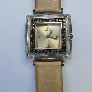 May include: A square-faced Swarovski watch with a gold-toned dial and hands. The watch features a clear, rectangular case with a gold-coloured bezel. The watch has a tan fabric strap.