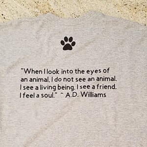 Puede incluir: Camiseta gris con una huella de pata y una cita sobre los animales: "Cuando miro a los ojos de un animal, no veo un animal. Veo un ser vivo. Veo un amigo. Siento un alma." ~ A.D. Williams