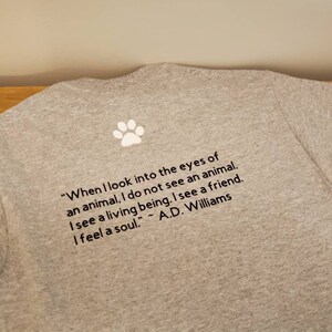 Puede incluir: Camiseta gris con una huella de pata blanca y una cita sobre los animales: "Cuando miro a los ojos de un animal, no veo un animal. Veo un ser vivo. Veo un amigo. Siento un alma." - A.D. Williams
