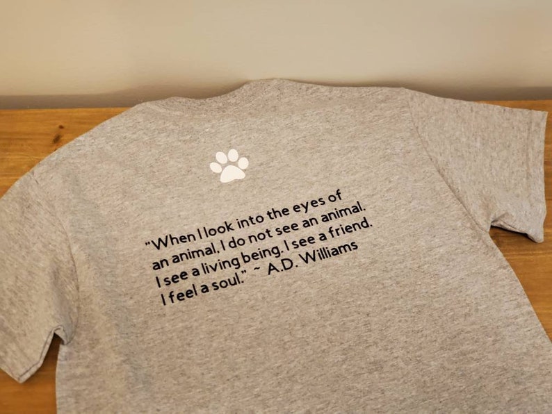 Puede incluir: Camiseta gris con una huella de pata blanca y una cita sobre los animales: "Cuando miro a los ojos de un animal, no veo un animal. Veo un ser vivo. Veo un amigo. Siento un alma." - A.D. Williams