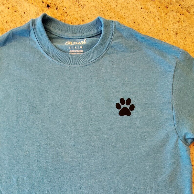 Puede incluir: Una camiseta azul claro con una huella de pata negra en el pecho izquierdo. La camiseta tiene una etiqueta Gildan en el cuello.