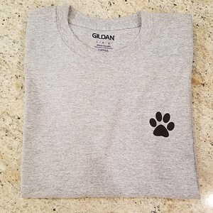 Puede incluir: Una camiseta gris con una huella de pata negra en el lado izquierdo. La camiseta tiene una etiqueta Gildan Lisle en la parte delantera.