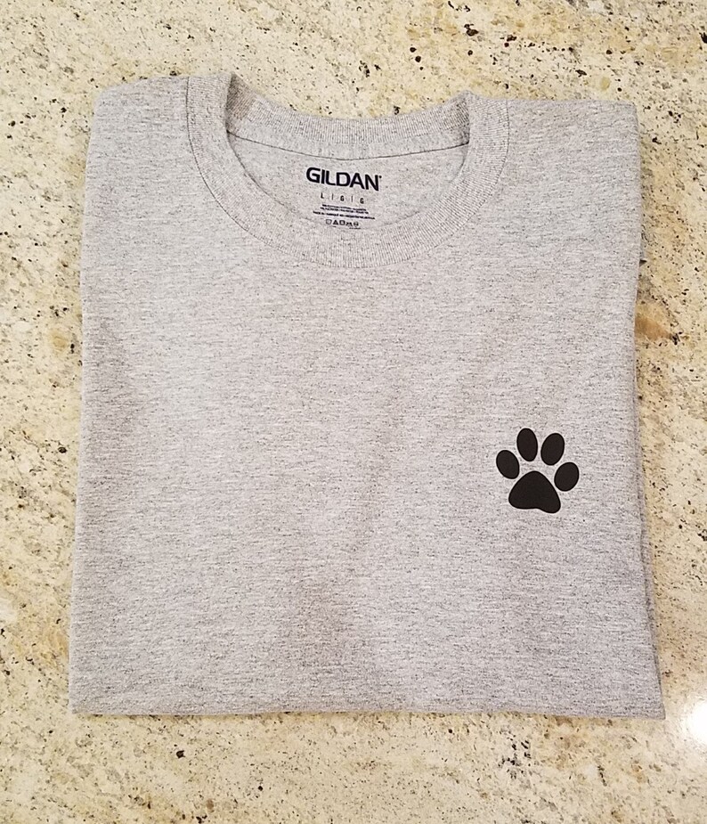 Puede incluir: Una camiseta gris con una huella de pata negra en el lado izquierdo. La camiseta tiene una etiqueta Gildan Lisle en la parte delantera.