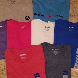Puede incluir: Ocho camisetas de la marca Gildan en varios colores, incluyendo gris, morado, rosa, blanco, azul, rojo y gris jaspeado. Las camisetas tienen una huella de pata, un coraz&oacute;n y el texto "Rescue. Repeat." impreso en ellas.