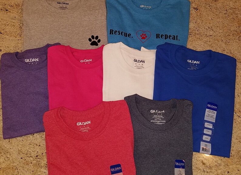 Puede incluir: Ocho camisetas de la marca Gildan en varios colores, incluyendo gris, morado, rosa, blanco, azul, rojo y gris jaspeado. Las camisetas tienen una huella de pata, un coraz&oacute;n y el texto "Rescue. Repeat." impreso en ellas.