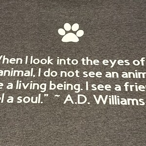 Puede incluir: Una camiseta gris con una huella de pata blanca y una cita sobre los animales: "Cuando miro a los ojos de un animal, no veo un animal. Veo un ser vivo. Veo un amigo. Siento un alma." - A.D. Williams