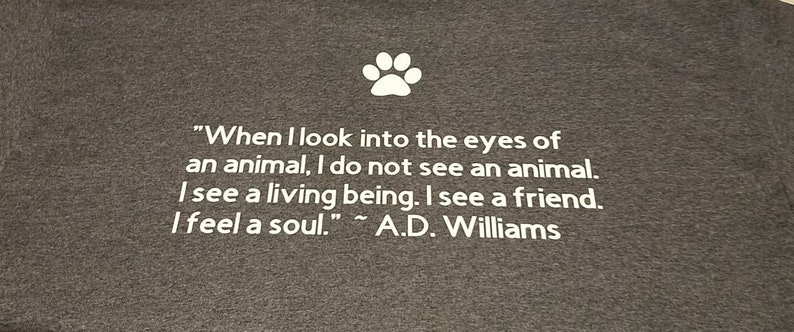 Puede incluir: Una camiseta gris con una huella de pata blanca y una cita sobre los animales: "Cuando miro a los ojos de un animal, no veo un animal. Veo un ser vivo. Veo un amigo. Siento un alma." - A.D. Williams